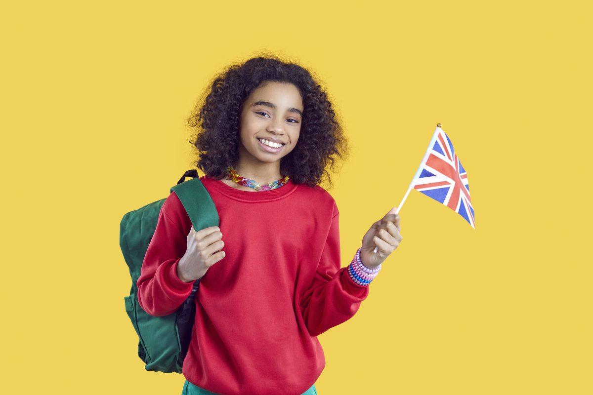 Learning foreign languages. Happy african american schoolgirl studying english posing with UK flag on yellow background. Ethnic preteen girl with backpack and flag of England smiling looking at camera