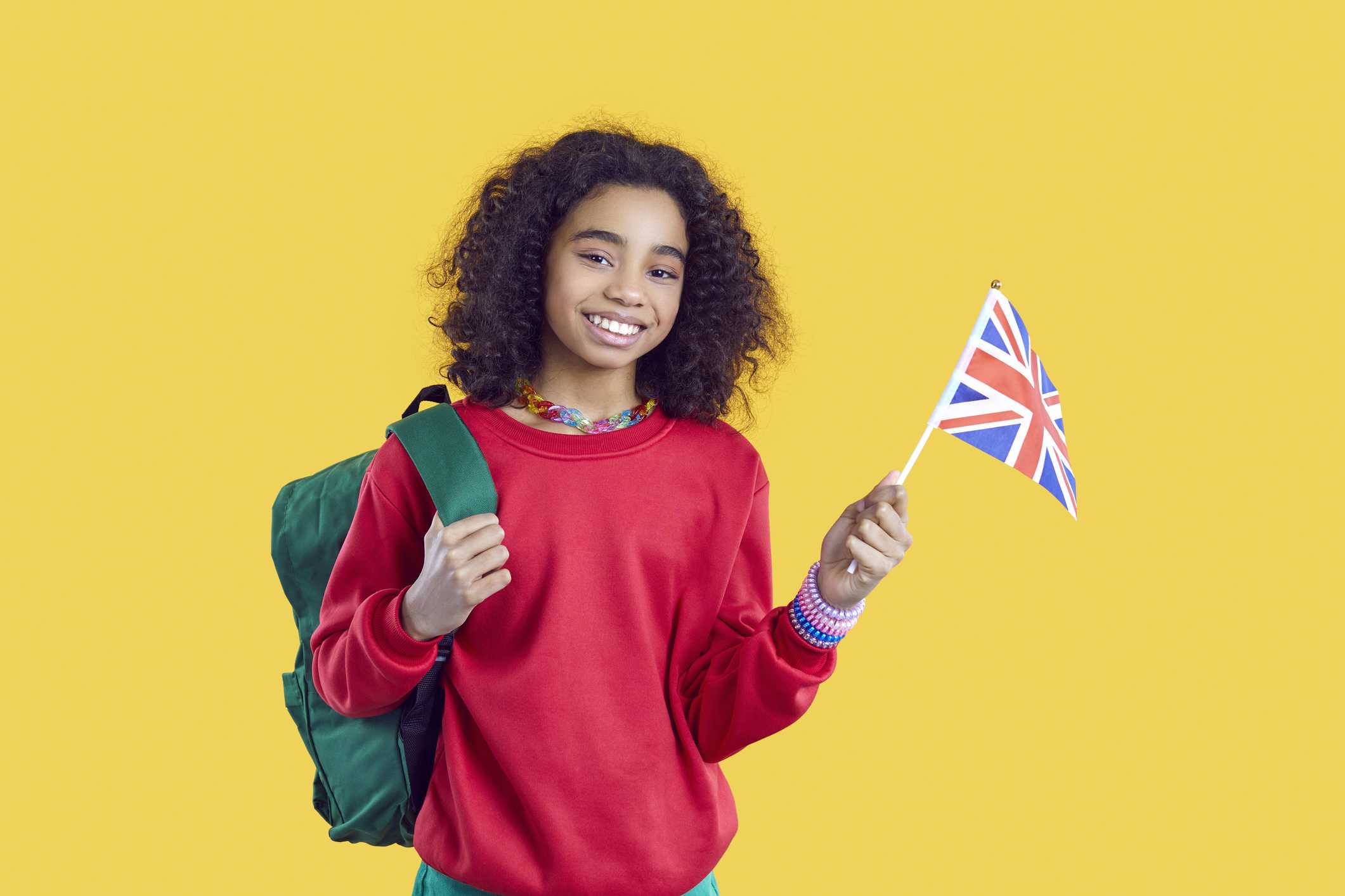 Learning foreign languages. Happy african american schoolgirl studying english posing with UK flag on yellow background. Ethnic preteen girl with backpack and flag of England smiling looking at camera